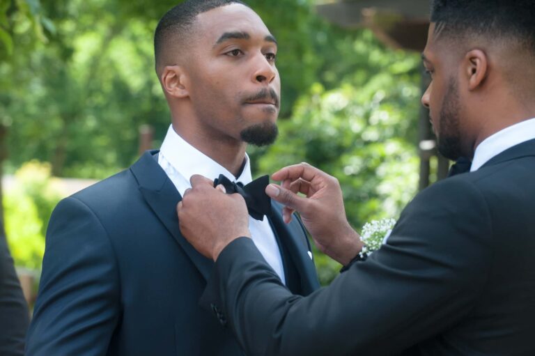 A groomsman straightens the groom's bow tie; Joelle and Tashan's wedding at Gilwell, Essex, Lovexposed Photography