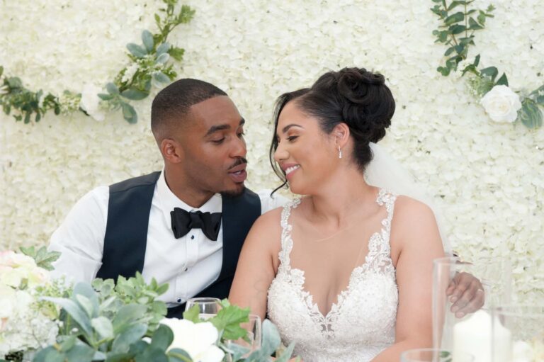 The couple at the top table in the marquee, framed by white flowers; Joelle and Tashan's wedding at Gilwell, Essex, Lovexposed Photography