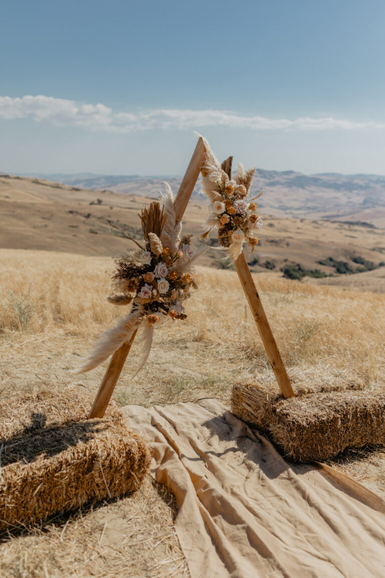 A wooden triangle arch with dried flowers and hay bales set for a ceremony in the wheat fields at Masseria Susafa, LOVIÙ Events