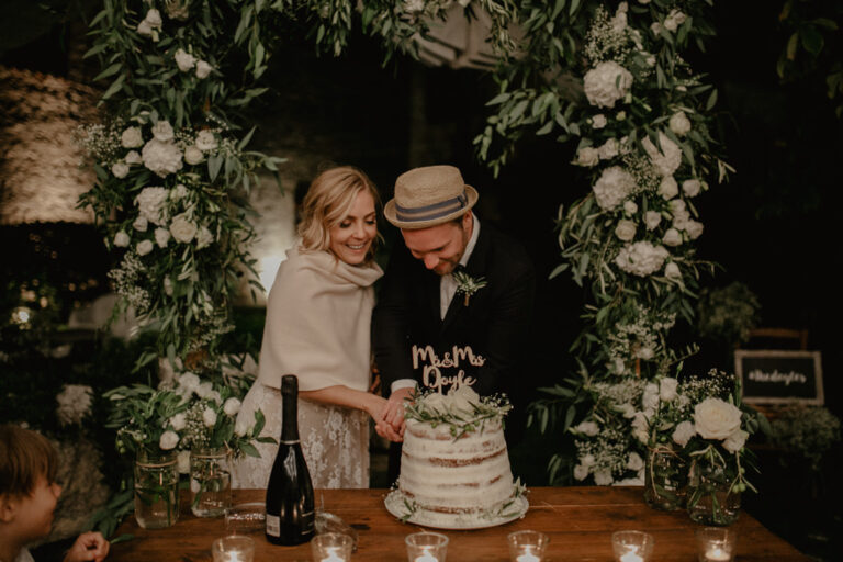 Cutting the cake beneath an arch of white flowers and greenery at Masseria Susafa, LOVIÙ Events