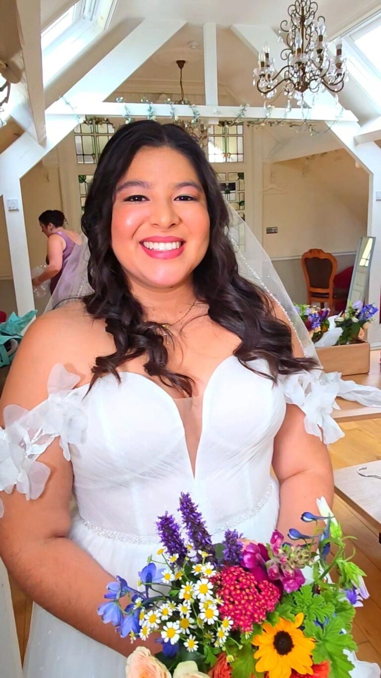 A bride with dark curls and a wildflower bouquet, in a wood-beamed room, Loxus Hair and Makeup
