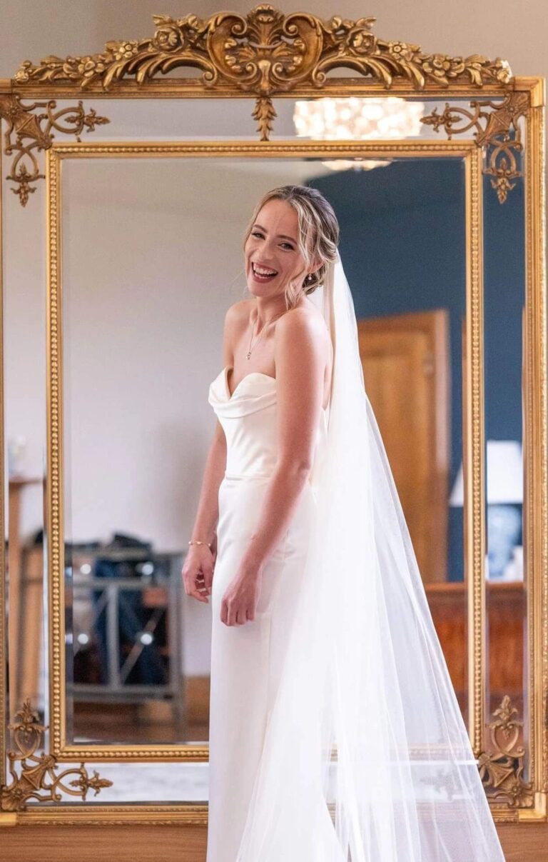 A bride in full length, veil trailing, reflected in an ornate gold mirror, Loxus Hair and Makeup