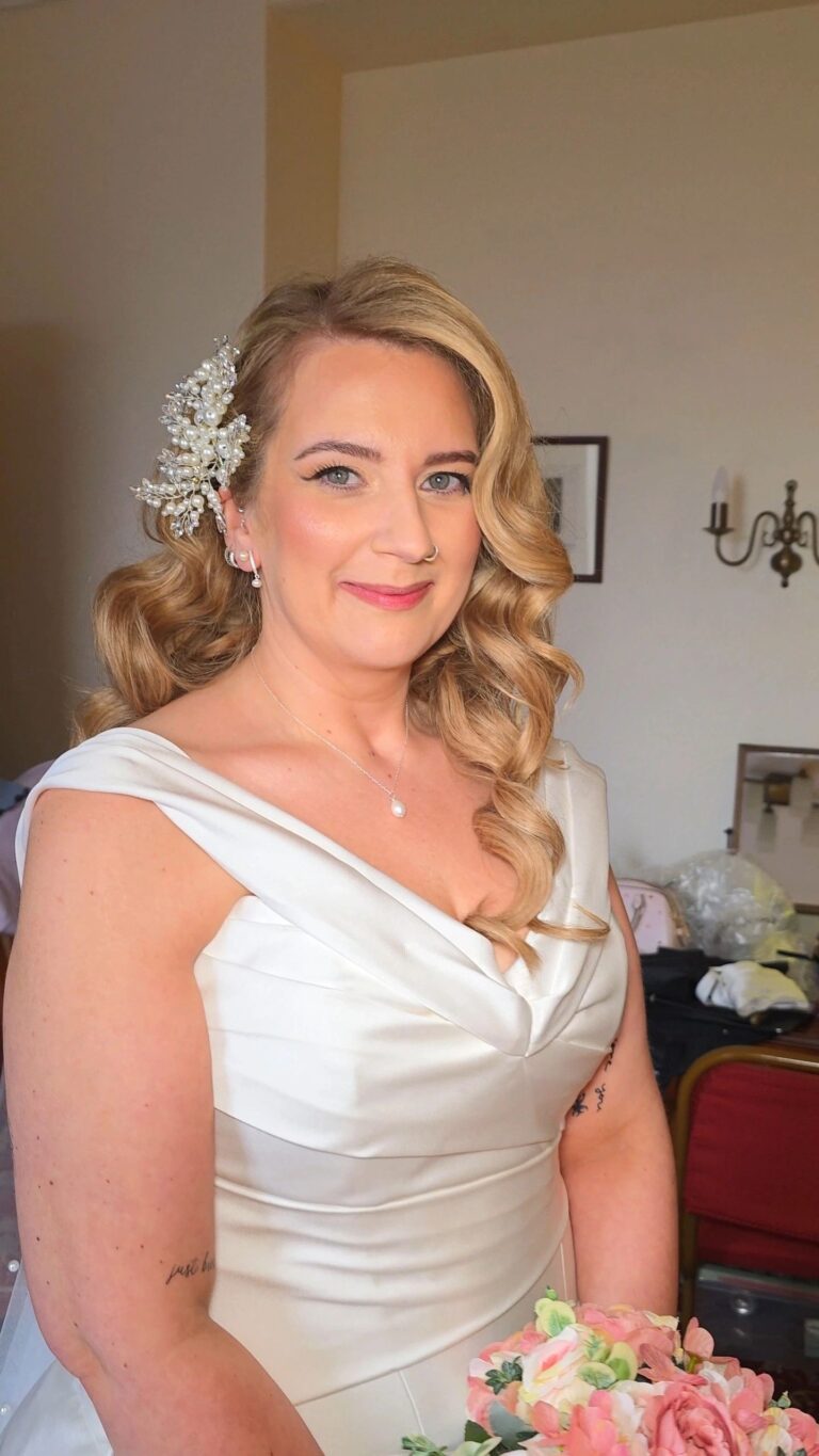 A bride's side-swept curls and floral hairpiece, a pearl necklace, Loxus Hair and Makeup