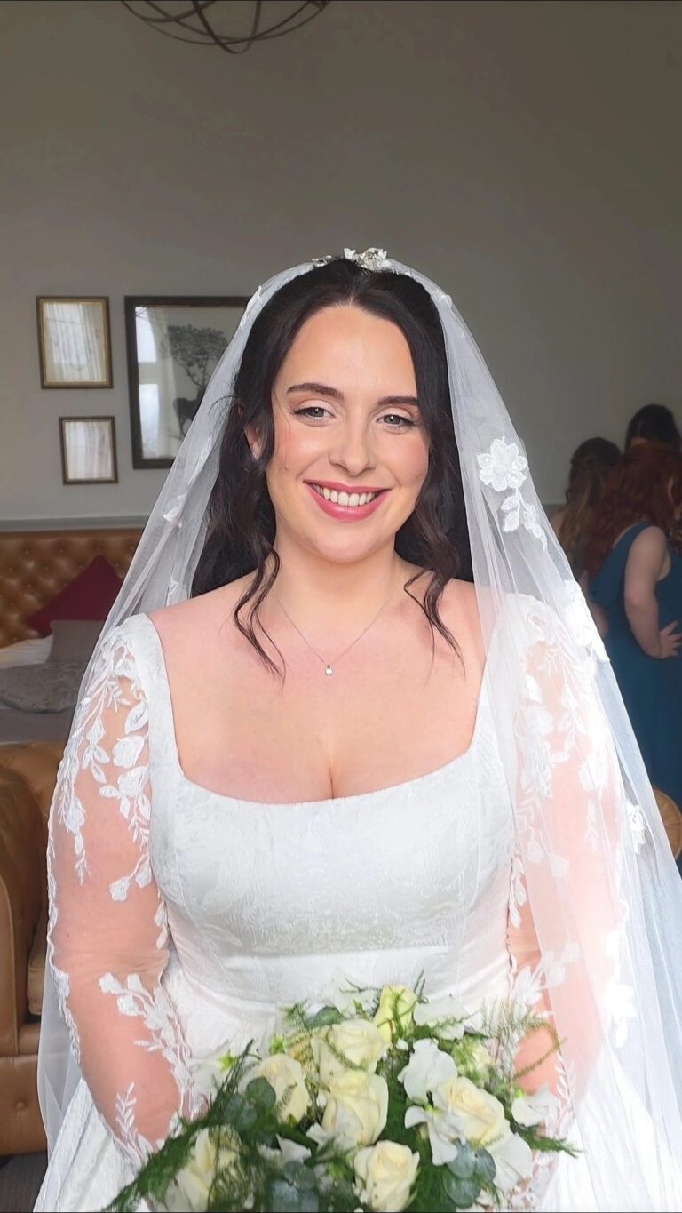A bride in a lace-sleeved gown and veil, bouquet in hand, family behind, Loxus Hair and Makeup