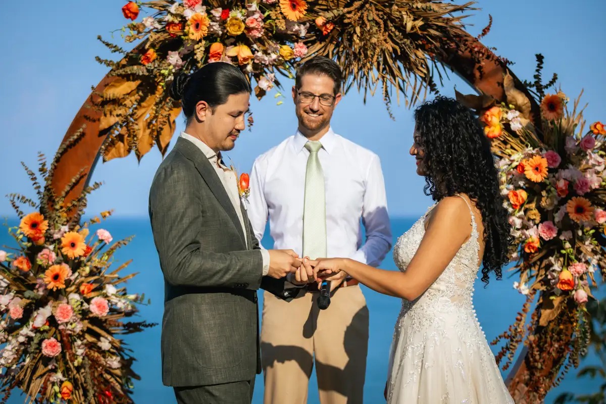 A couple exchanging rings beneath a round arch of dried and fresh flowers above the sea, Cape Panwa, Phuket, Lucida Image