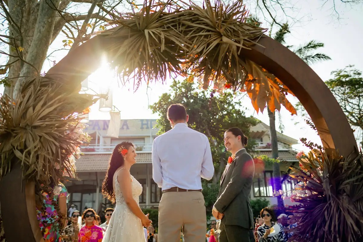 Sunlight breaking through a floral arch as the couple stand with their celebrant, Cape Panwa, Phuket, Lucida Image