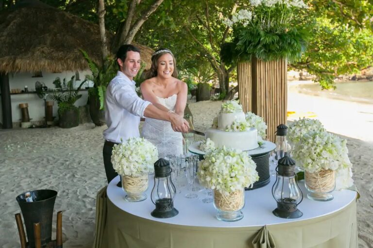A couple cutting a white wedding cake on a table set with white flowers and lanterns, Rayavadee, Krabi, Lucida Image