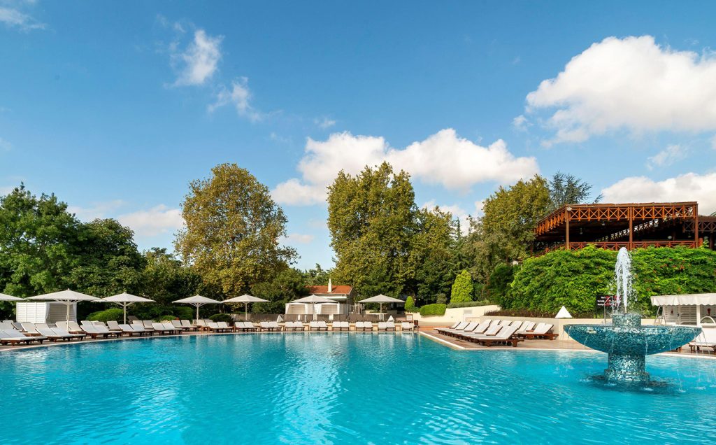 A swimming pool with a fountain, sun loungers and parasols among the hotel gardens