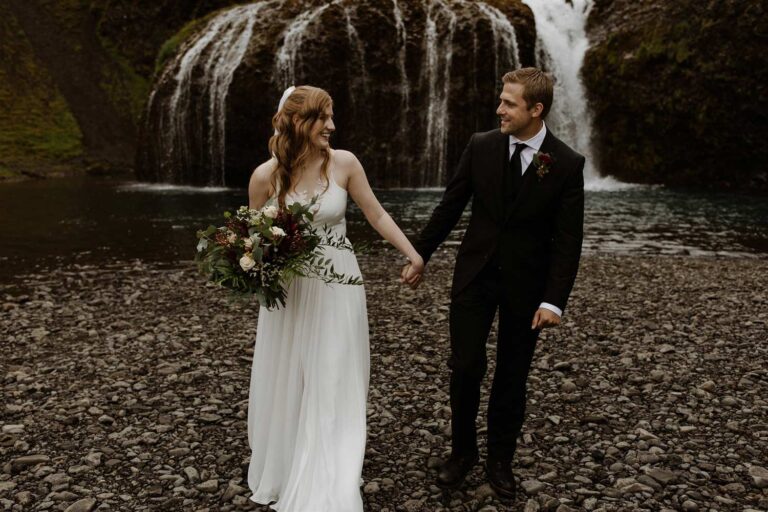 A bride and groom hold hands in front of a waterfall with a trailing green bouquet, Luxwedding Iceland
