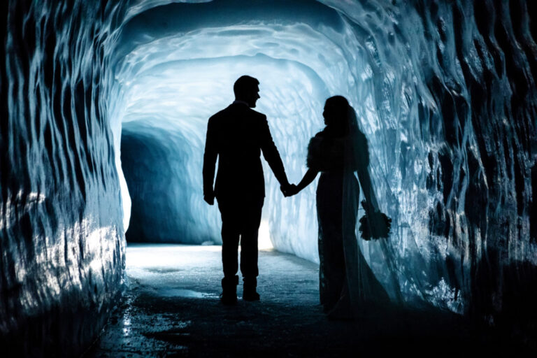 A couple hold hands in silhouette at the mouth of a deep blue ice tunnel, Luxwedding Iceland