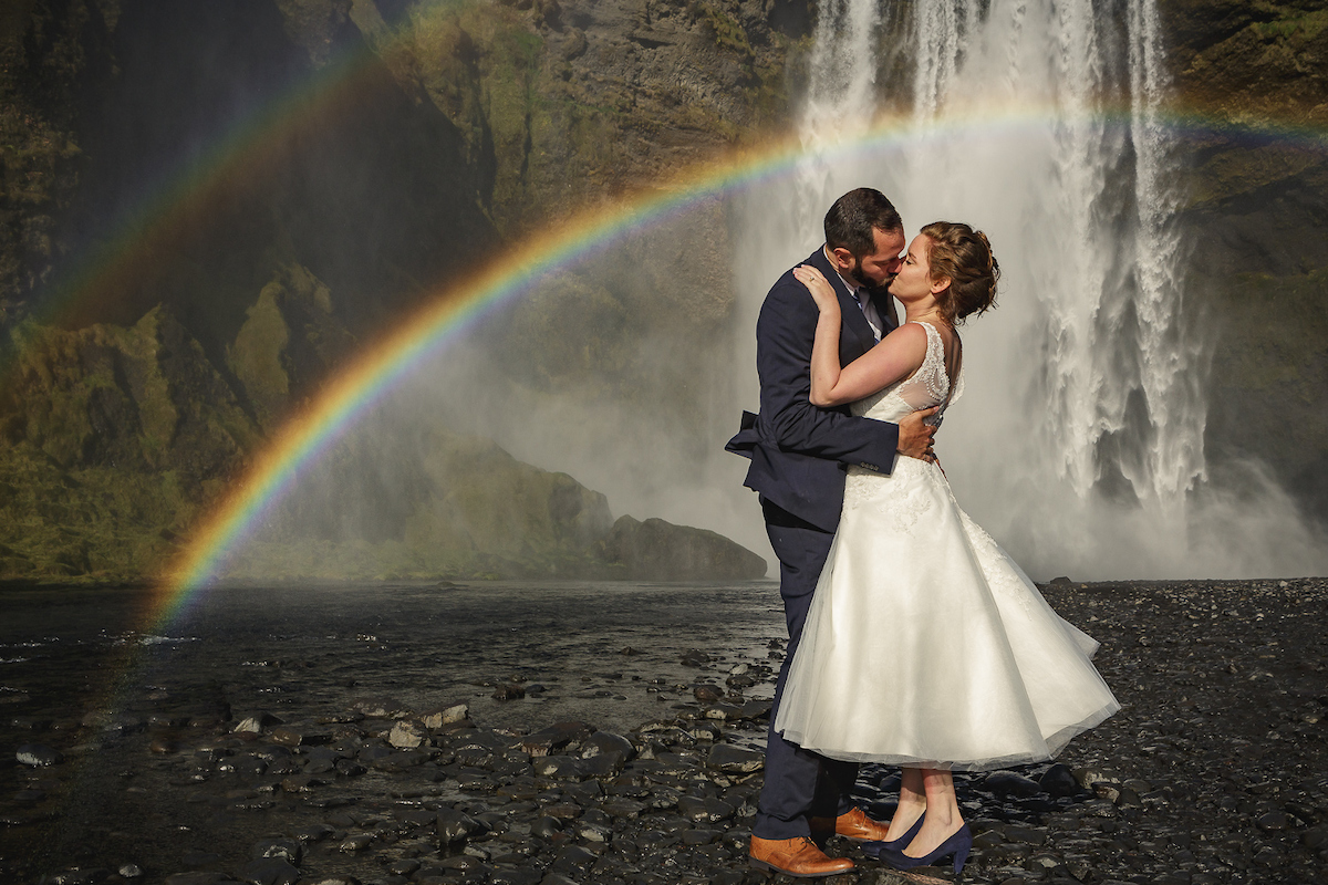A couple embrace beneath a waterfall with a rainbow arching over the black pebble shore, Luxwedding Iceland