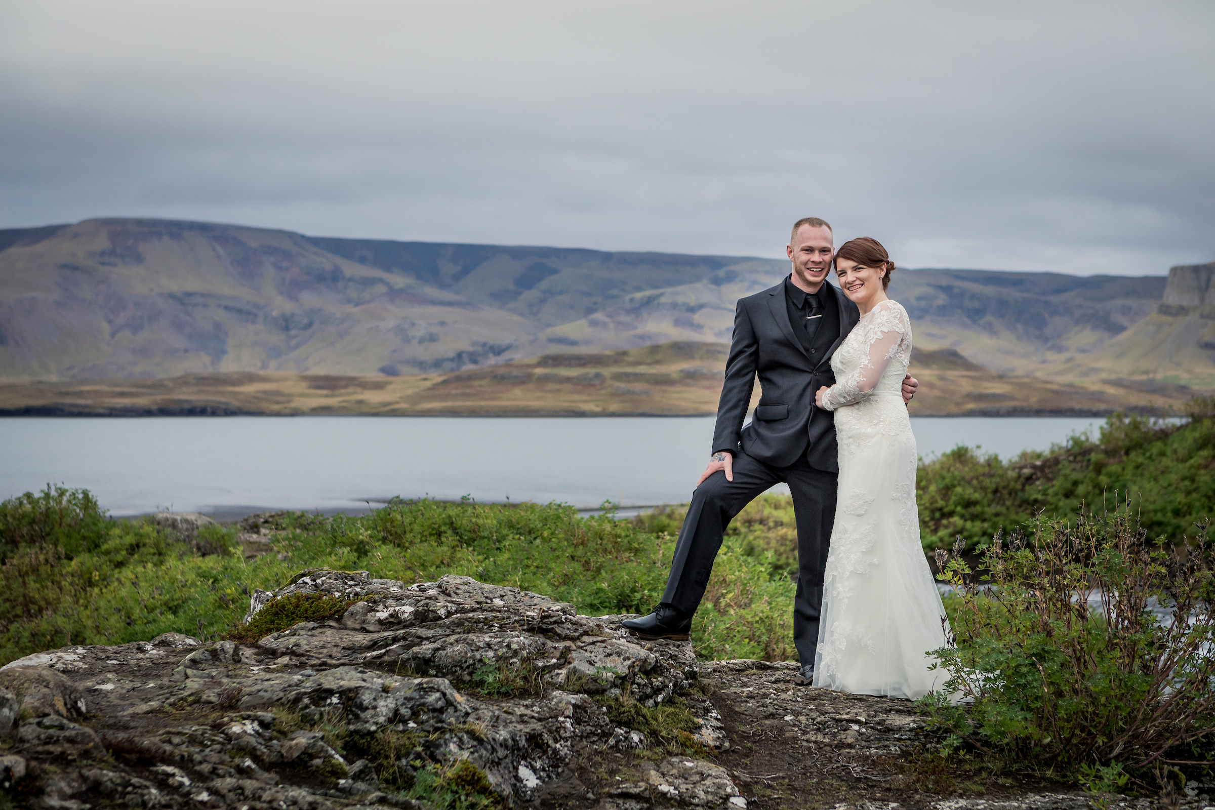 A couple pose on a rocky outcrop above a lake with mountains behind, Luxwedding Iceland