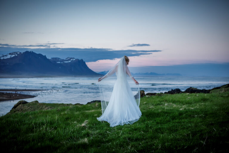 A bride lifts her veil on a green headland beside the sea and mountains, Luxwedding Iceland
