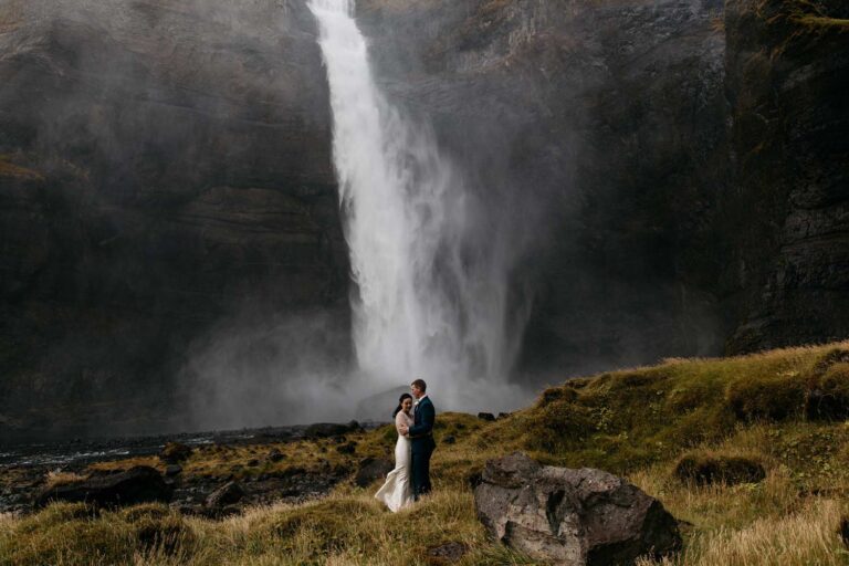 A couple stand below a tall waterfall in an autumn valley, Luxwedding Iceland