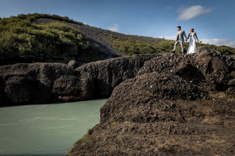 A couple hold hands on dark lava rock above a turquoise glacial river, Luxwedding Iceland