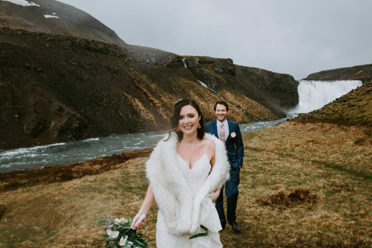 A bride in a white fur stole and her groom beside a river leading to a waterfall, Luxwedding Iceland