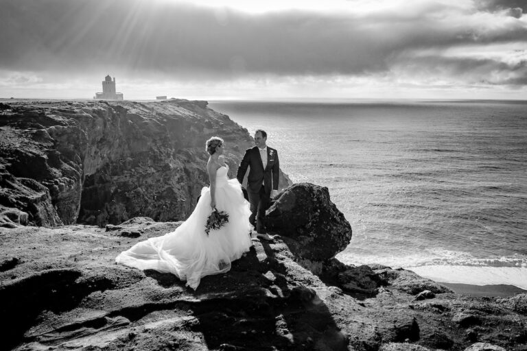 A couple on a sea cliff in black and white, with a lighthouse on the headland beyond, Luxwedding Iceland