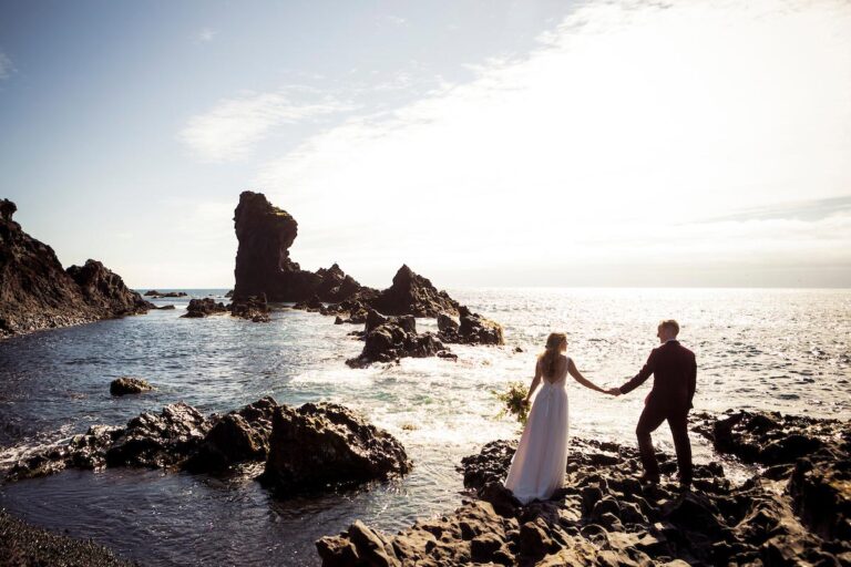 A couple hold hands on a rocky shore facing the sea at golden hour, Luxwedding Iceland