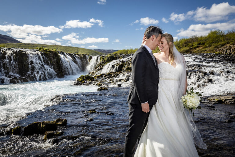 A couple embrace in front of a wide, low waterfall under a blue sky, Luxwedding Iceland