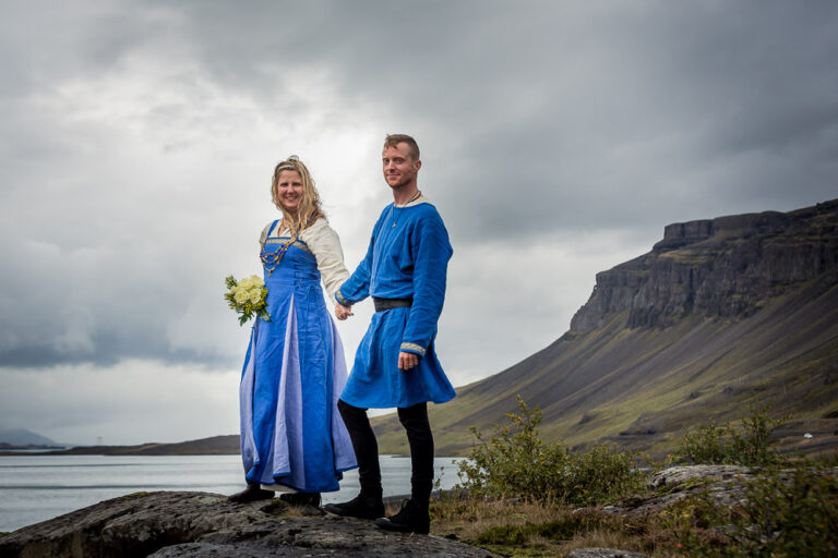 A couple in blue, traditional-style wedding attire hold hands on a rock above a fjord, Luxwedding Iceland