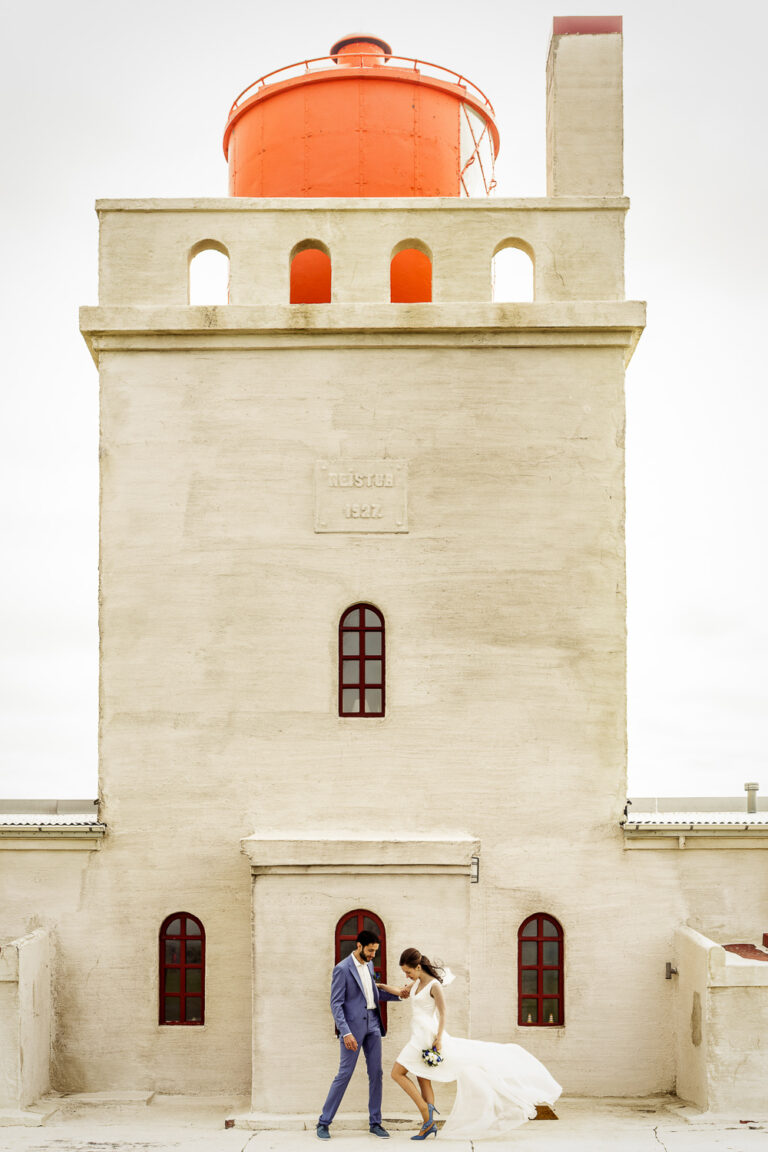 A couple walk past a white lighthouse tower with an orange lantern room, Luxwedding Iceland