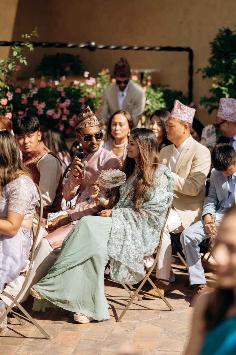 Guests seated in colour beneath the flower arch, topi caps among them, Magnolia Wedding Planner