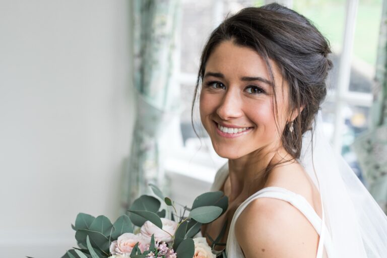 A bride in a veil, holding her bouquet, smiling in a garden, Makeup By Becky Flynn