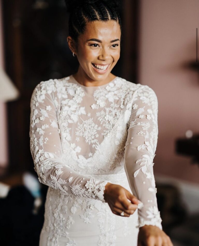 A bride buttoning a long-sleeved lace gown, a tall window behind her, Makeup By Becky Flynn