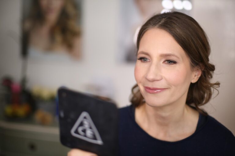A woman checking her makeup in a hand mirror, framed bridal portraits on the wall behind her, Makeup By Becky Flynn