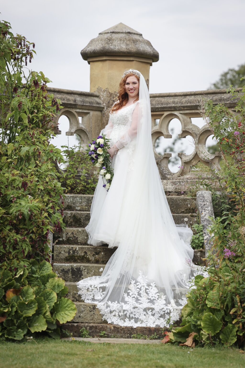 A bride alone on stone garden steps, cathedral veil trailing behind her, Makeup By Mirna