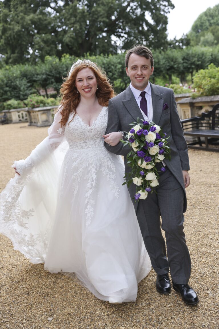 A bride and groom walking arm in arm, gravel path, bouquet of purple and white, Makeup By Mirna