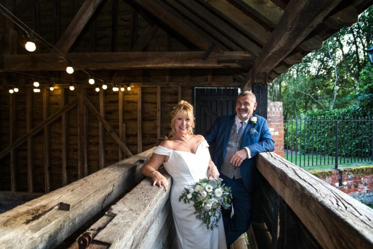 A bride and groom in a timber barn, festoon lights strung overhead, Makeup By Mirna