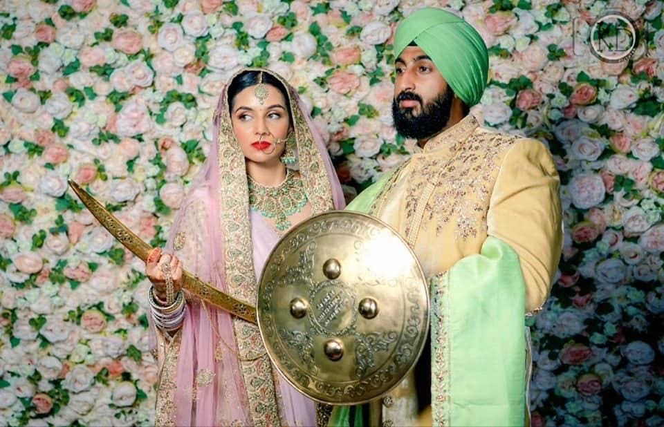 A Sikh bride and groom, ceremonial sword and shield, a floral backdrop, Makeup By Mirna