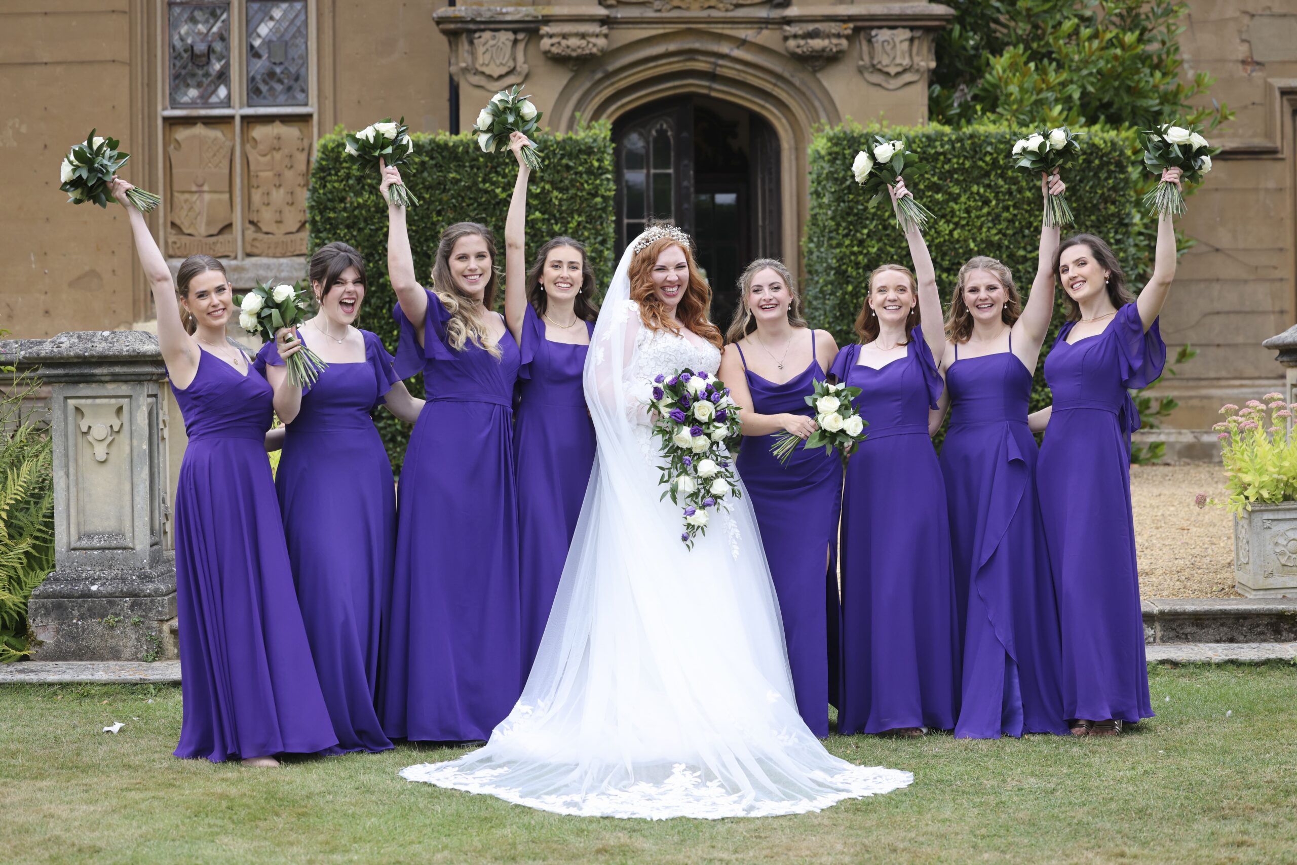 A bride and eight bridesmaids in violet, bouquets raised, a country house behind, Makeup By Mirna