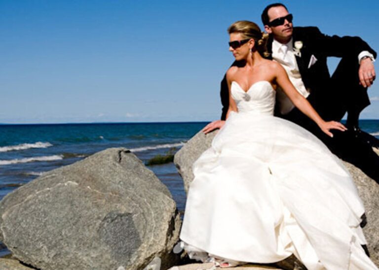 A couple seated on lakeshore boulders, the bride in a full-skirted gown, Maple Leaf Weddings