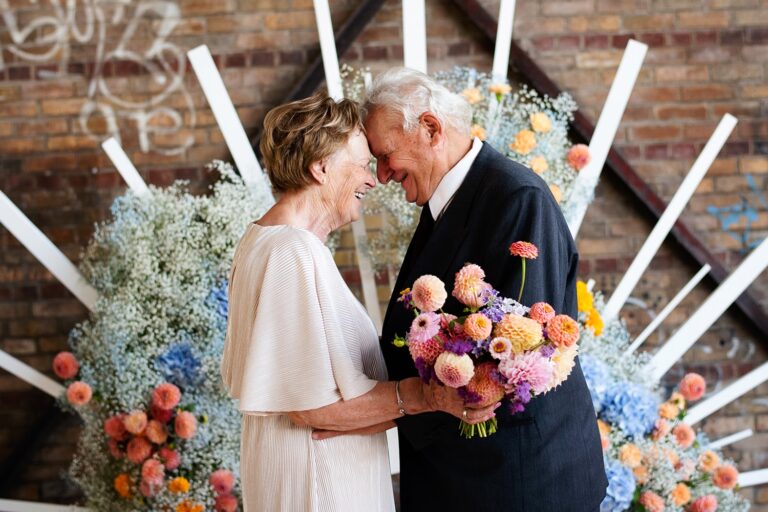 An older couple with foreheads together, surrounded by pastel flowers and a brick wall, Maple Leaf Weddings