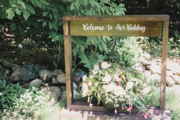 A wooden welcome sign reading "Welcome to Our Wedding" set among ferns and rocks in a garden, Maple Leaf Weddings