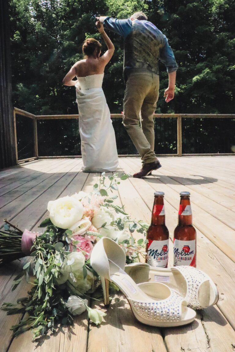 A bride and groom crossing a deck, bridal shoes, beer bottles and a bouquet in the foreground, Maple Leaf Weddings
