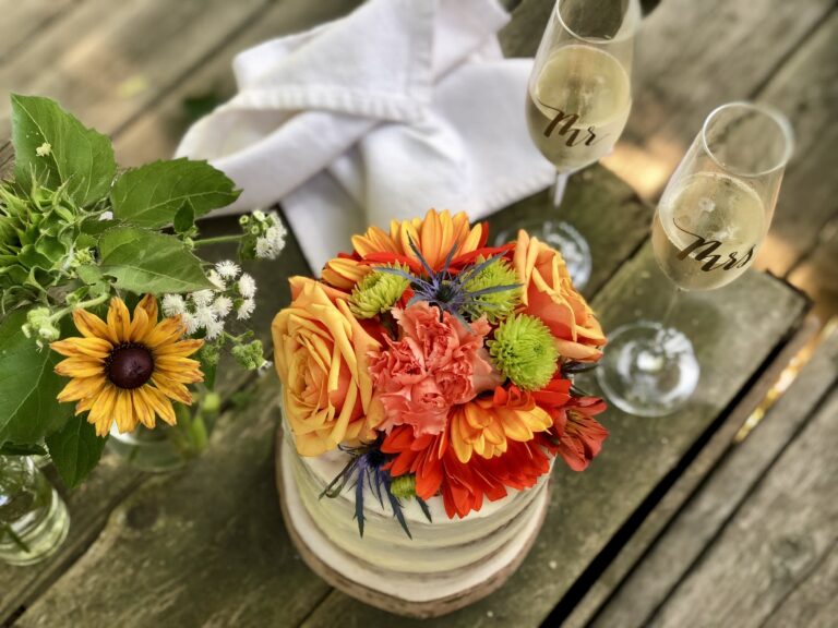 A bright posy of orange roses and sunflowers on a rustic table with two champagne flutes, Maple Leaf Weddings