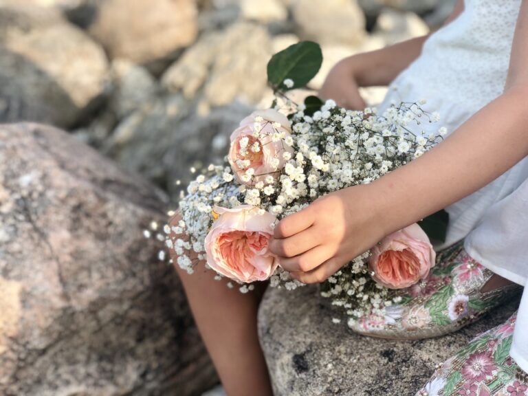 A flower girl holding a posy of blush roses and gypsophila on a lakeshore rock, Maple Leaf Weddings