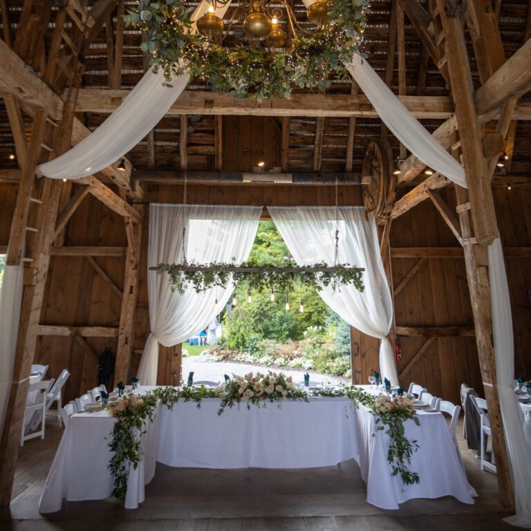 A barn set for a wedding, with white drapes, a hanging floral installation and a long head table, Maple Leaf Weddings