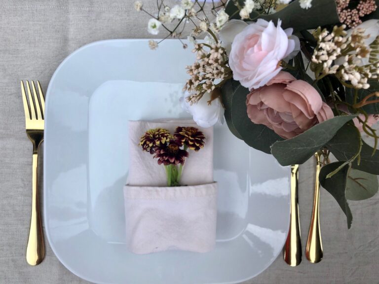 A place setting with gold cutlery, a napkin tucked with a small posy and a blush floral arrangement, Maple Leaf Weddings
