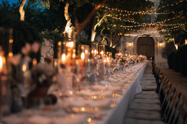 A long table lit by hundreds of candles under trees, running to an arched doorway, Marica Events