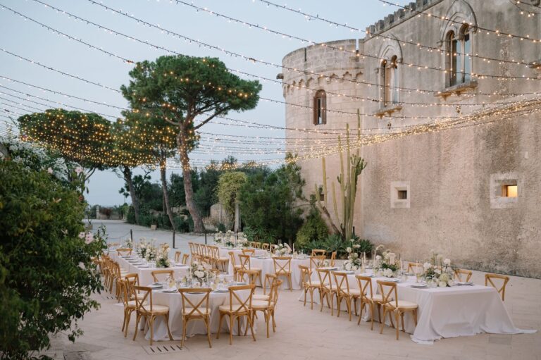 Round tables in a castle courtyard at dusk, festoon lights strung between the pines, Marica Events