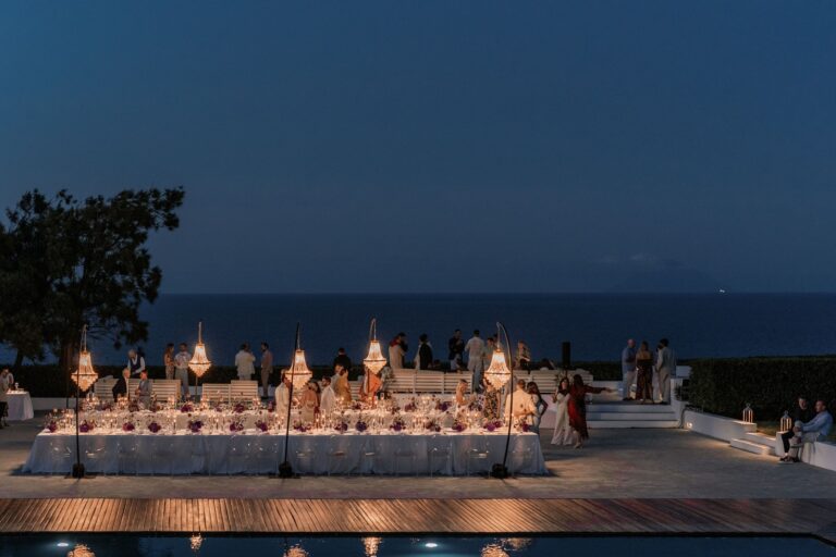 A long white table on a terrace above the sea at blue hour, lanterns down its length, Marica Events