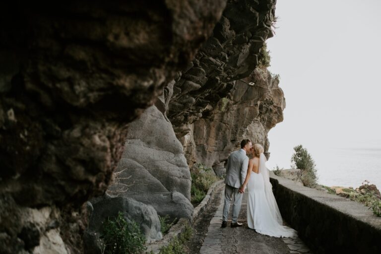 The couple on a stone path beneath a great overhanging rock face, the sea beyond, Marica Events