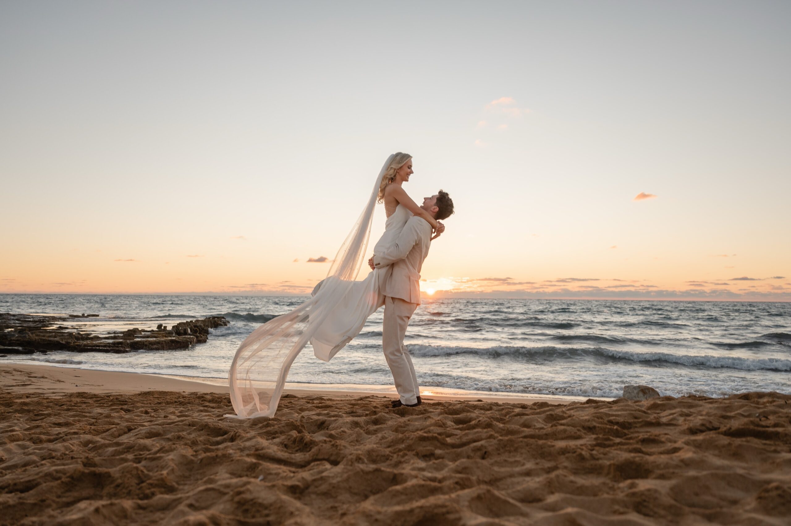 Groom lifting his bride on the sand at sunset, veil trailing in the breeze, Paphos coast, Marry Me Cyprus