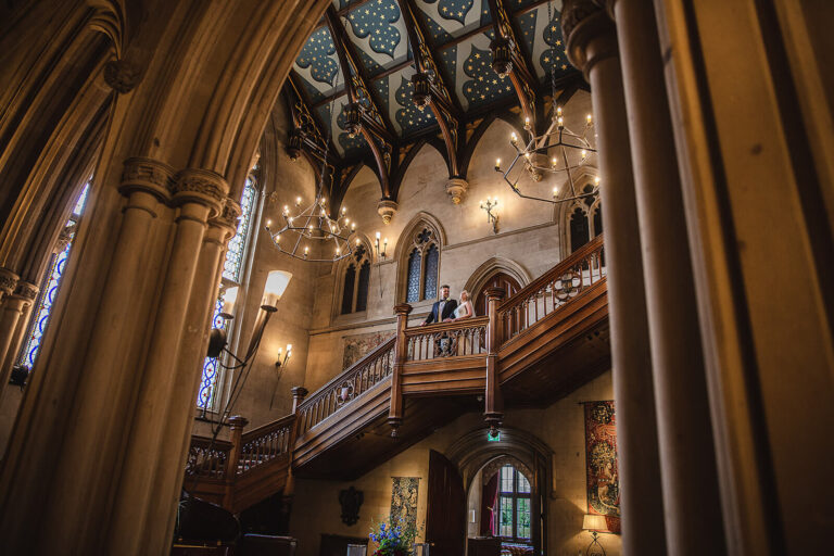 Looking up at the Great Hall's oak staircase, gallery and starred painted ceiling, lit by chandeliers, Matfen Hall