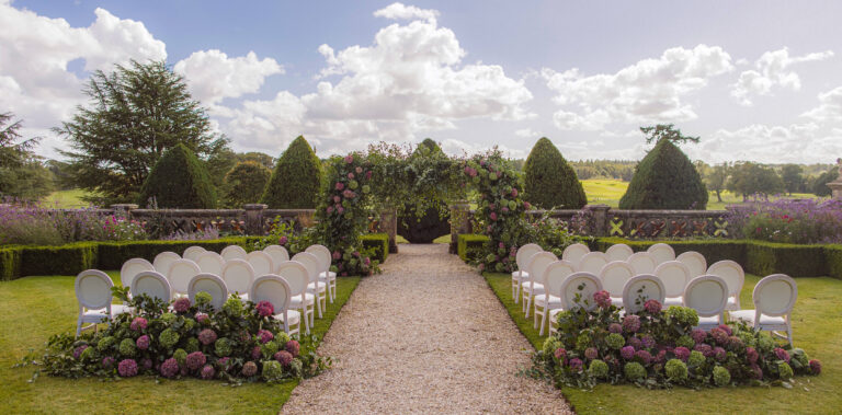Rows of white chairs set for an outdoor ceremony either side of a gravel aisle in Le Jardin Hollandais, with clipped yews and a rose arch, Matfen Hall
