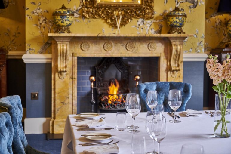 The Sapphire Room's marble fireplace with a lit fire beside a laid dining table, Matfen Hall